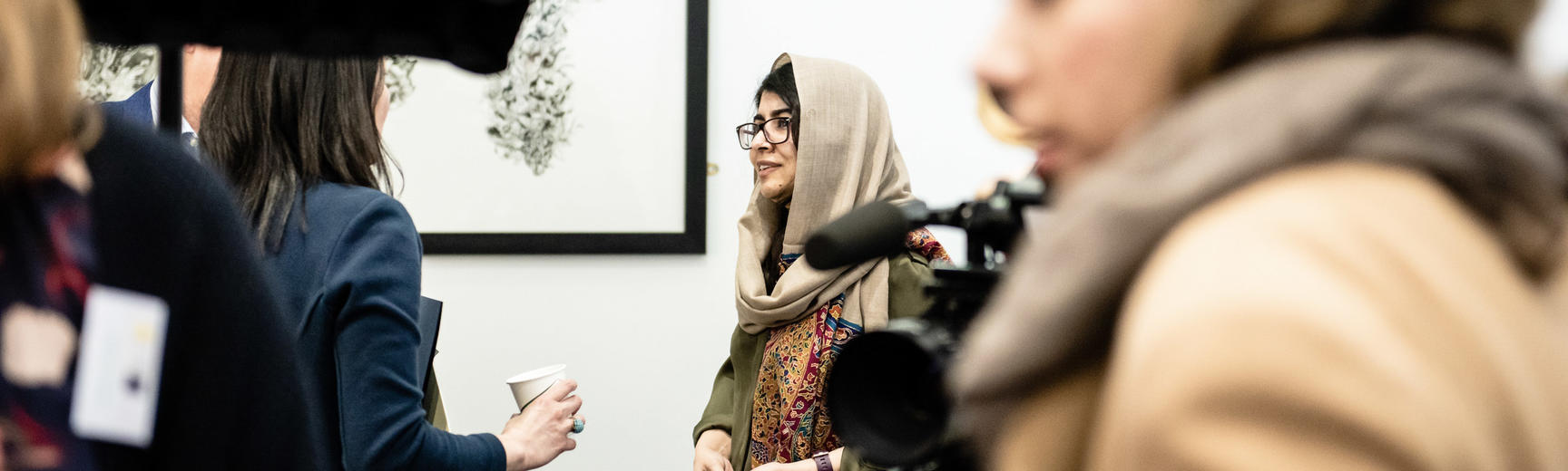 Malala Yousafzai speaks with a young woman in the background, in the foreground media equipment